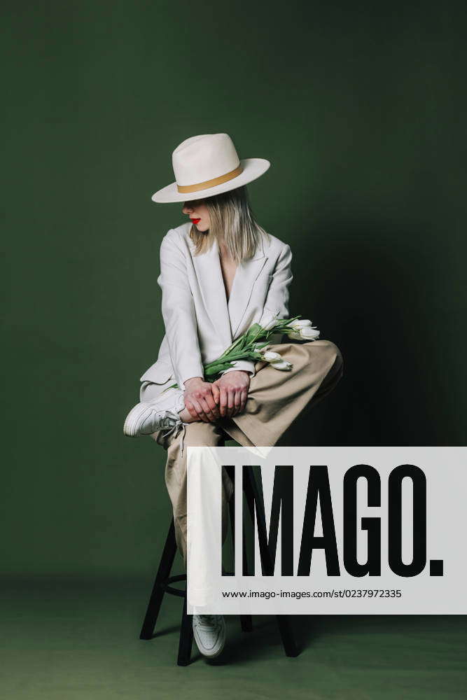 Fashionable Woman With Flowers Wearing Hat On Stool In Front Of Green