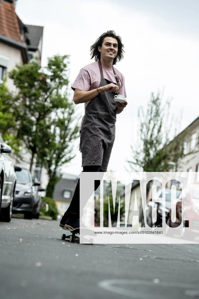 Smiling male barista holding coffee cup while skateboarding on road in city model released Symbolfoto KNSF08169