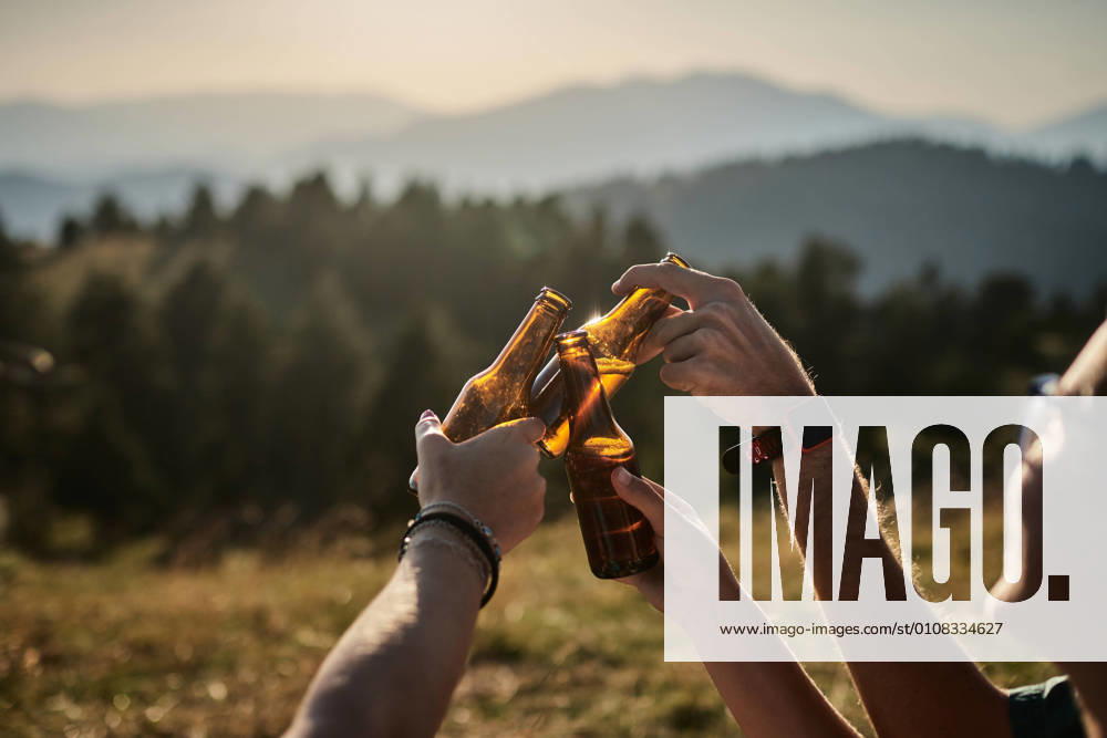 Company of unrecognizable friends relaxing in highland area and clinking bottles of beer while enjoying sunset in summer during weekend
