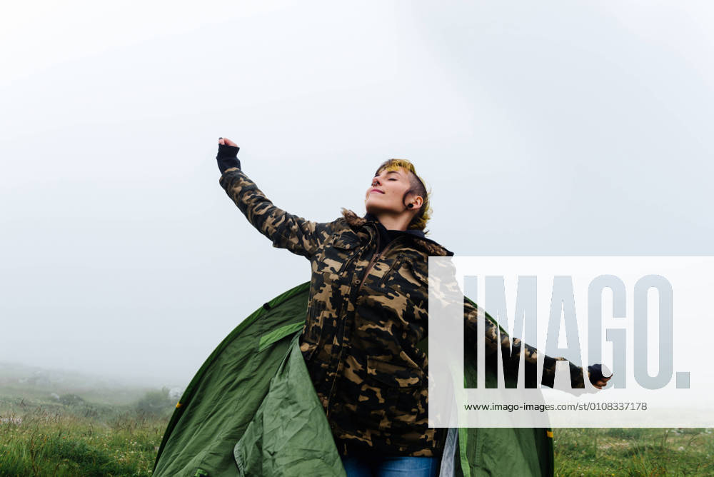 IMAGO Creative: Klimawandel - Nachhaltiges Reisen Positive female in warm clothes getting out of camping tent and stretching body during foggy morning in nature, Model released