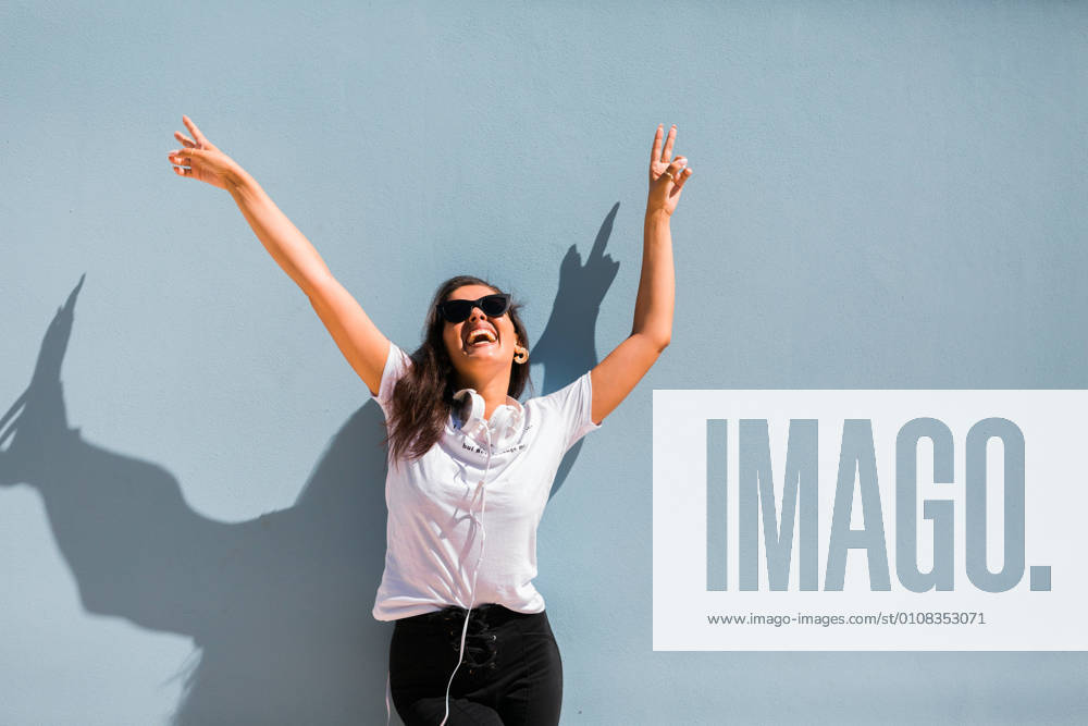 Carefree beautiful woman in casual outfit and headphones on neck standing with hands up beside blue wall of building on city street, Model released