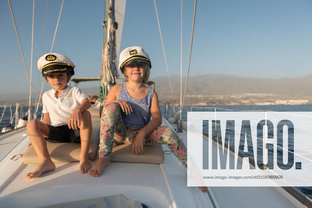 Positive kids in captain hats sitting on deck of expensive boat floating on water in sunny day, Model released, Property released
