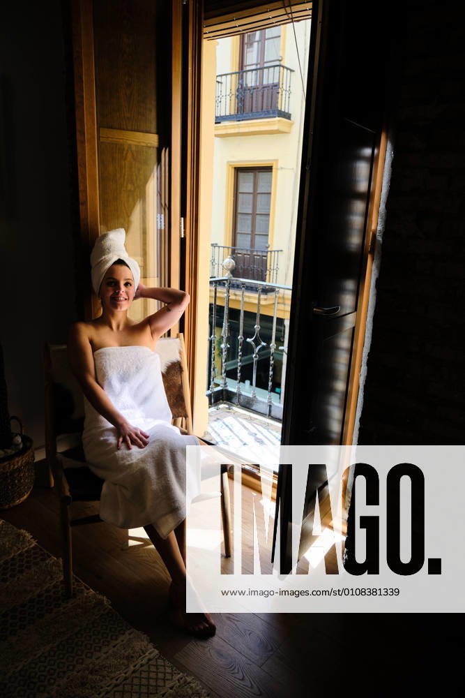 IMAGO Creative, Jahreszeit Sommer, Hitze IMAGO Creative, Sommer, Schatten-Pl?tze bei Hitze Serene relaxed happy female in white towel on body and head sitting on chair by the balcony door in apartment with wooden floor on sunny day and looking away