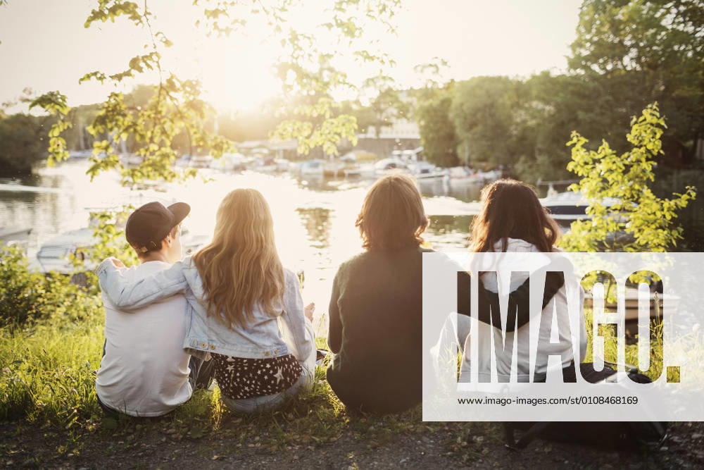 IMAGO Creative: Sommer, Sommertage im Stadtpark IMAGO Creative, Jahreszeit Sommer, Sommer in der Stadt Rear view of teenagers sitting with arms around at lakeshore , model released