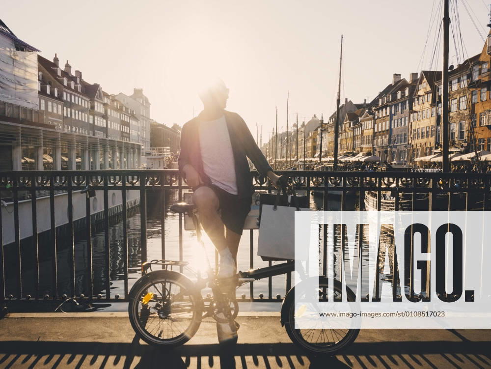 IMAGO Creative, Jahreszeit Sommer, Sommer in der Stadt Full length back lit of man standing with bicycle and shopping bags on footbridge in city , model released