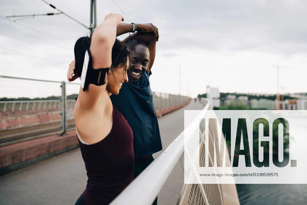 IMAGO Creative, Jahreszeit Sommer, Sommer in der Stadt Smiling male and female athletes stretching hands on footbridge over sea against sky , model released