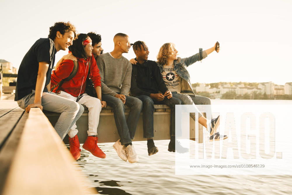 IMAGO Creative, Jahreszeit Sommer, Sommer in der Stadt Happy male friends taking selfie while sitting side by side on pier by river , model released