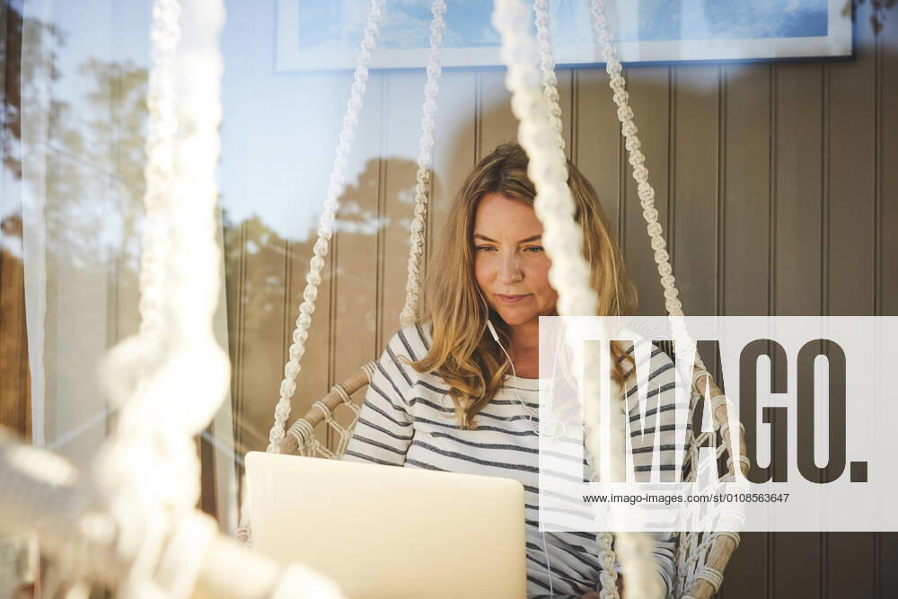 Mid adult female with headphones working over laptop while sitting in rope swing , model released, property released