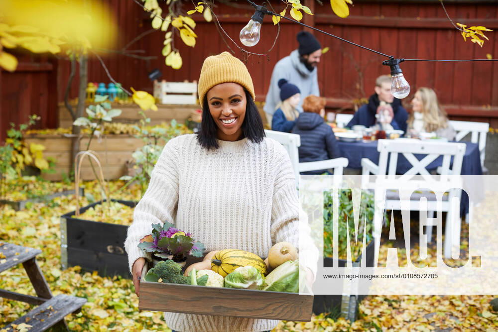 IMAGO Creative: Landleben - Essen Portrait of happy woman standing with vegetable basket while family and friends sitting at table in yard , model released, property released