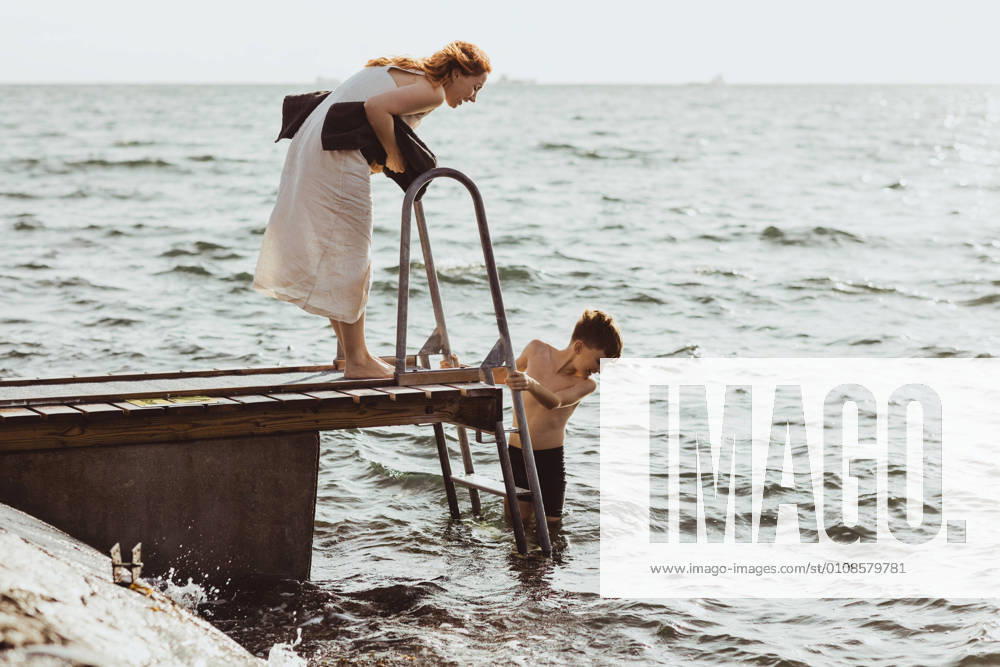 IMAGO Creative, Jahreszeit Sommer, Sommerferien Side view of woman guiding while son moving down on ladder in sea Sweden, Gothenburg, model released
