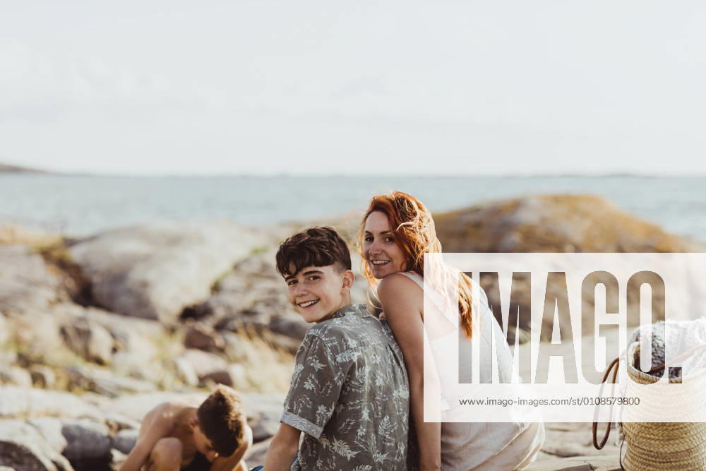 IMAGO Creative, Jahreszeit Sommer, Sommerferien Portrait of smiling mother and son bonding while sitting over rocky land during sunny day Sweden, Gothenburg, model released