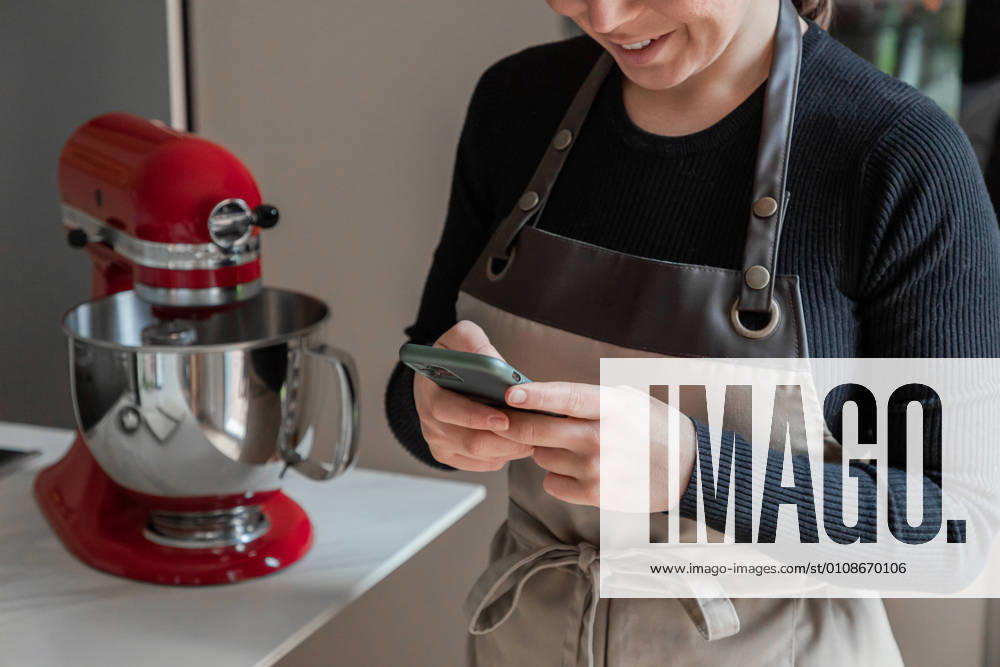 IMAGO Creative: Haushaltsger?te - K?chenmaschine Crop positive smiling housewife in apron using mobile phone and reading recipe while standing next to table with red stand mixer in home kitchen