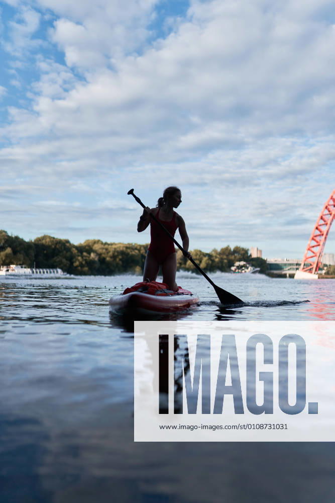 IMAGO Creative, Jahreszeit Sommer, Sommer in der Stadt Front view of woman standing on paddle board in river and rowing with paddle under bridge in city