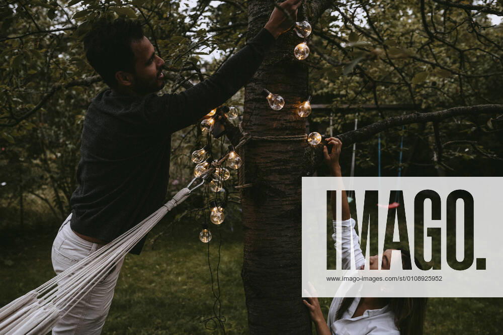 Male and female friends hanging lighting equipment in backyard during dinner party Sweden, Stockholm, model released, property released