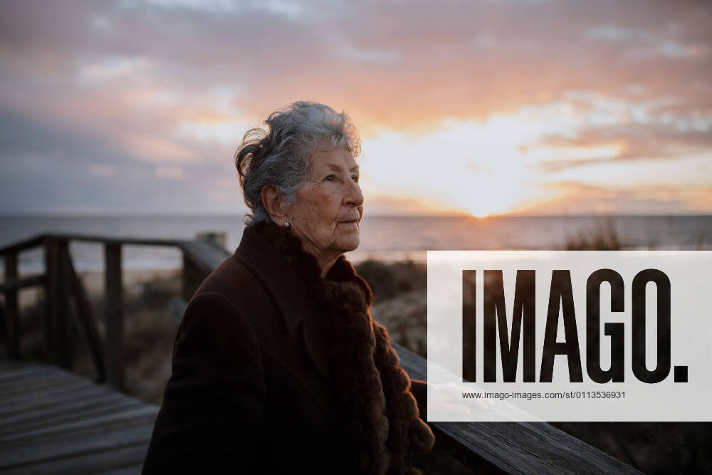Side view of elderly female traveler in casual clothes standing on wooden pier on sandy beach and enjoying seascape at sunset