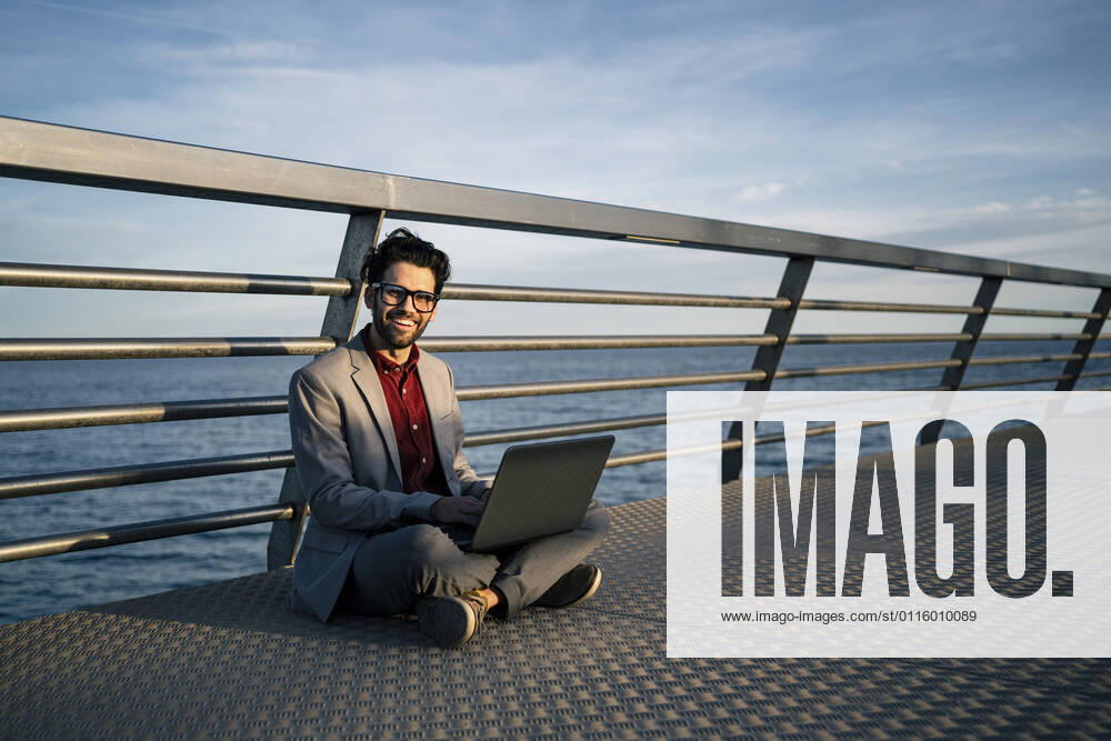 Smiling freelance worker with laptop sitting cross legged on pier model released Symbolfoto RCPF00834