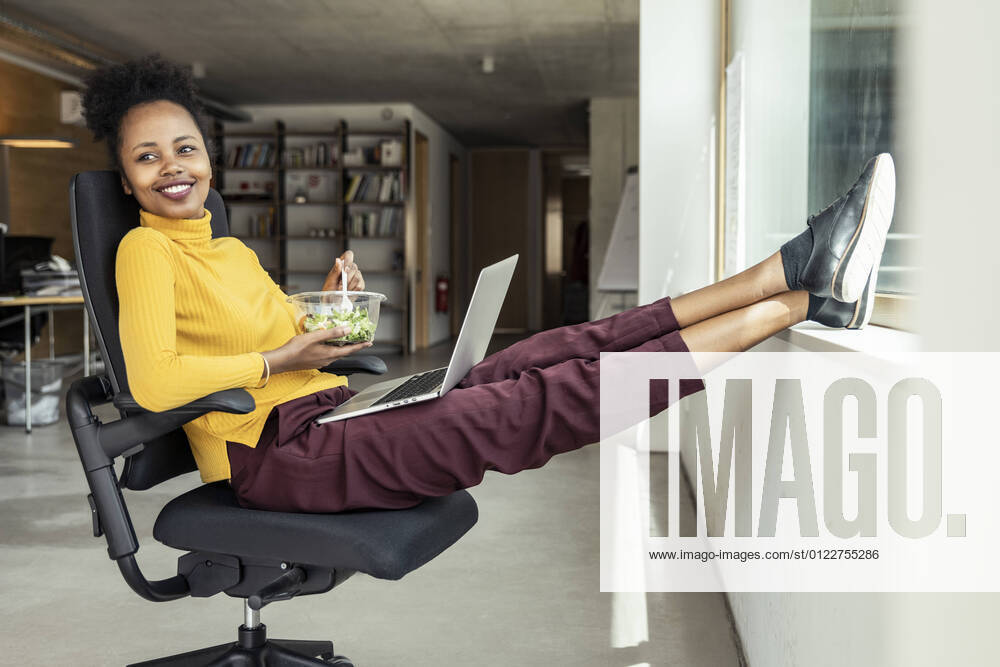 Smiling businesswoman with laptop looking away while having salad on chair at apartment model released Symbolfoto property released UUF23339