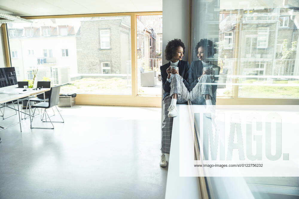 Businesswoman holding coffee cup while sitting on window sill at office model released Symbolfoto property released UUF23526