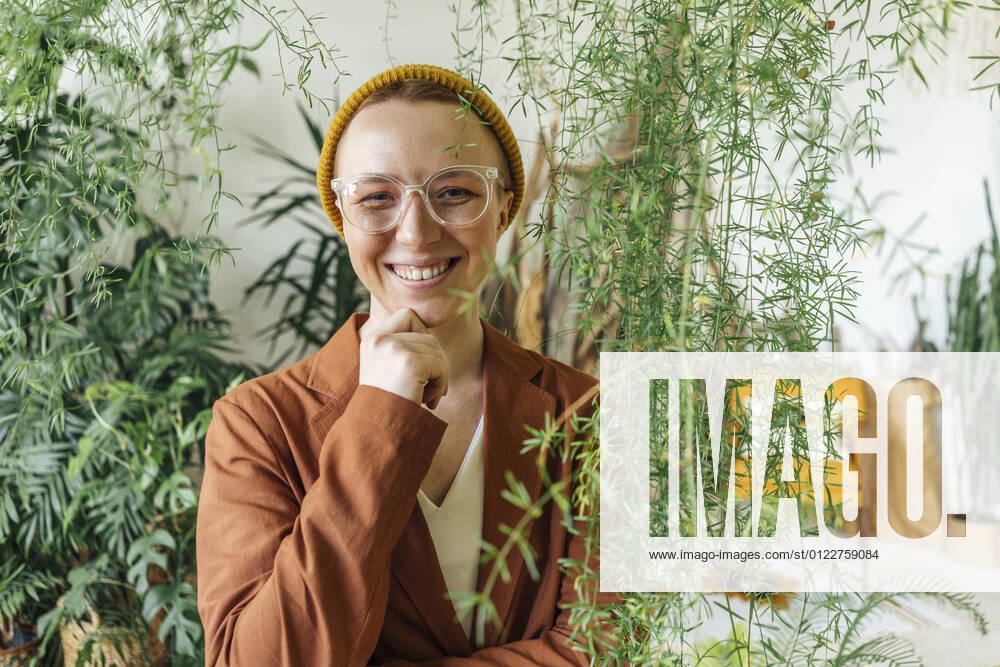 Smiling female entrepreneur with hand on chin standing by plants at work place model released Symbolfoto property released VPIF04136