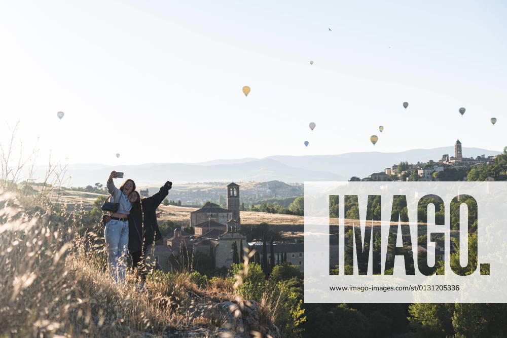 IMAGO Creative, Jahreszeit Sommer, Sommer in der Stadt Spain, Castile and Leon, Segovia, Two sisters taking selfie with flying hot air balloons and historical city buildings model released Symbolfoto JAQF00693