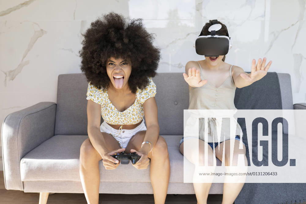 Afro woman sticking out tongue while female friend wearing virtual reality headset at home model released Symbolfoto property released JCCMF03236