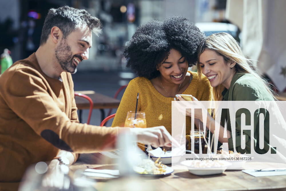 IMAGO Creative, Food, Tapas Happy female friends sharing mobile phone by male friend at restaurant model released Symbolfoto property released JSRF01606