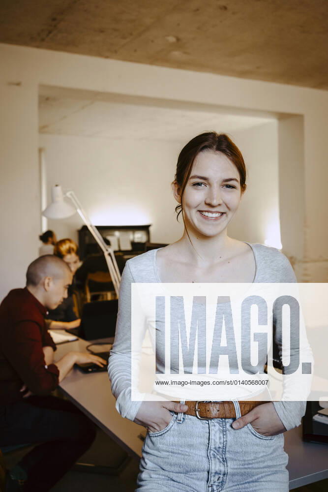 Portrait of smiling businesswoman with hands in pockets while colleague working at desk in office Germany, Berlin, model released, property released