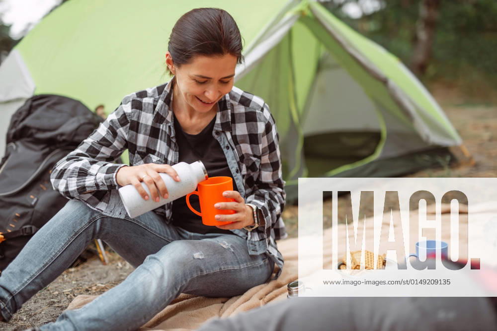 IMAGO Creative: Sommer, In den Bergen Camping in mountains forest Beautiful adult girl drinking tea in camping in forest with tent on background and smiling. Young woman traveller during hiking in mountains