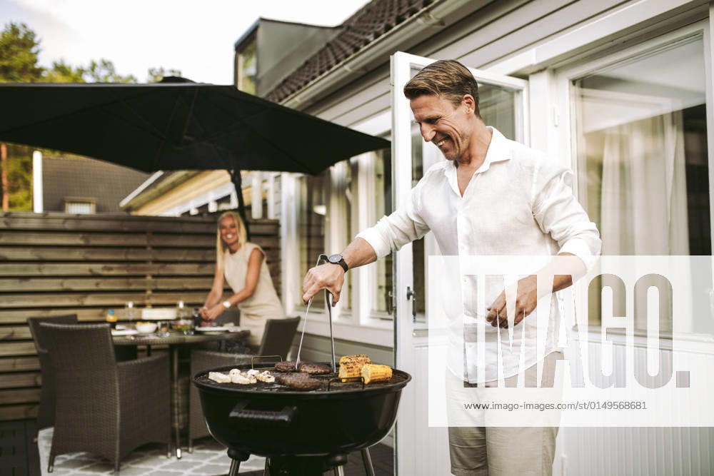 Smiling mature man preparing barbecue meal while in woman in background at back yard Sweden, Gothenburg, model released, property released