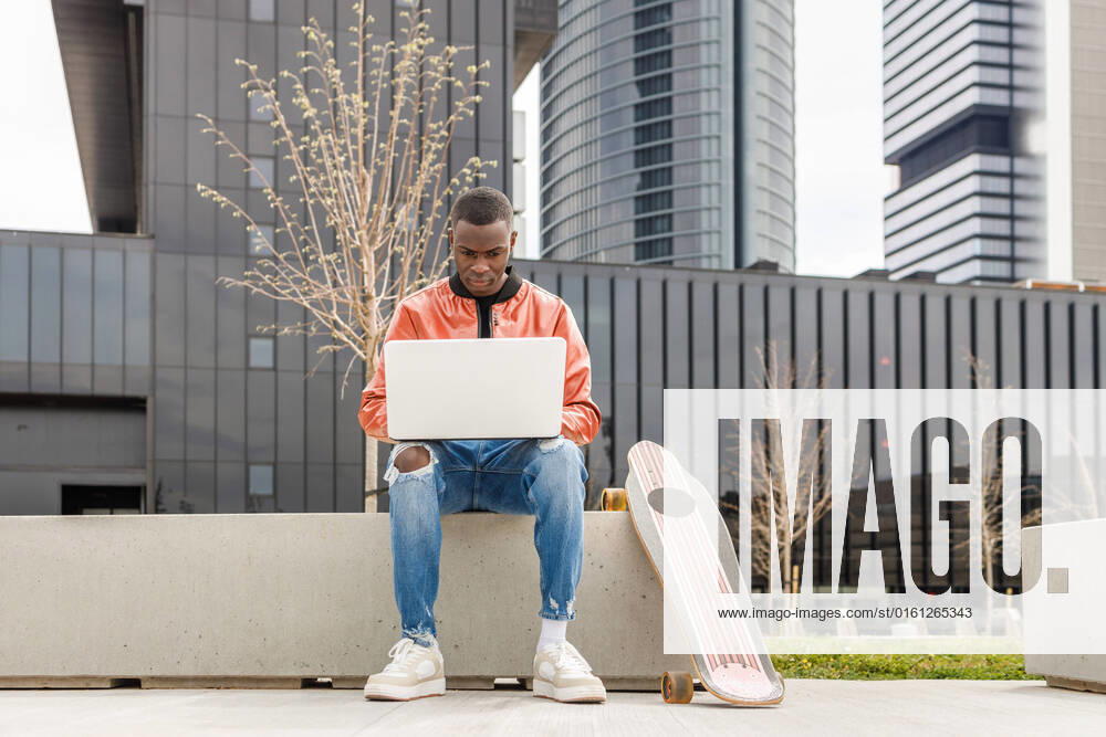 IMAGO Creative: Business - Work-Life Balance Concentrated young African American male freelancer in trendy outfit sitting on stone bench and working remotely on laptop near longboard in city park, Model released IgnacioFerrandiz_ZGenerationStudent_5.jpg