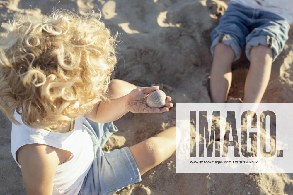 IMAGO Creative, Jahreszeit Sommer, Sommerferien From above of unrecognizable little child with curly blond hair in casual clothes holding pebble while sitting on sandy beach and playing with crop sibling during summer holidays SinuheGorris_EveningWithFriends_00034.jpg
