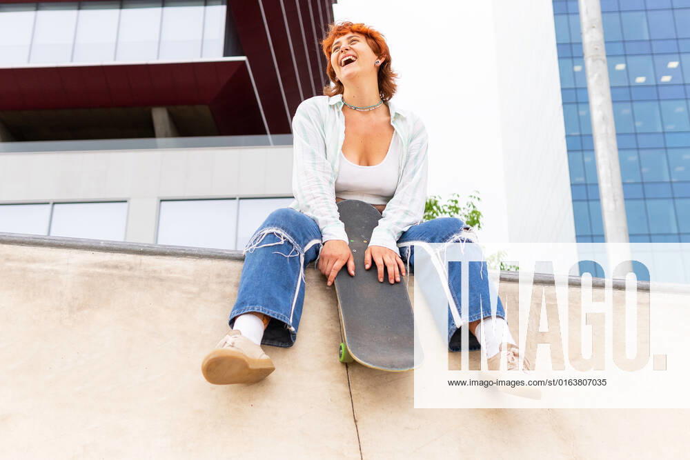 IMAGO Creative: Sommer, Sommertage im Stadtpark Full body of content female in casual wear with skateboard looking away while sitting on concrete border with skate park in city, Model released DavidMunoz_NatiAndHugoSkaters_00017.jpg