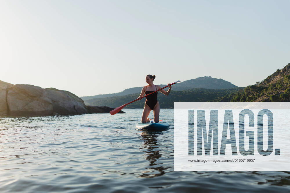 IMAGO Creative: 2022 Sport Favoriten IMAGO Creative: Ende des Sommers Full body of active female in swimwear floating on paddleboard in rippling lake against rock and shore with forest on summer day, Model released MiguelPalomo_WomanWithPaddleSurfInLake_11.jpg