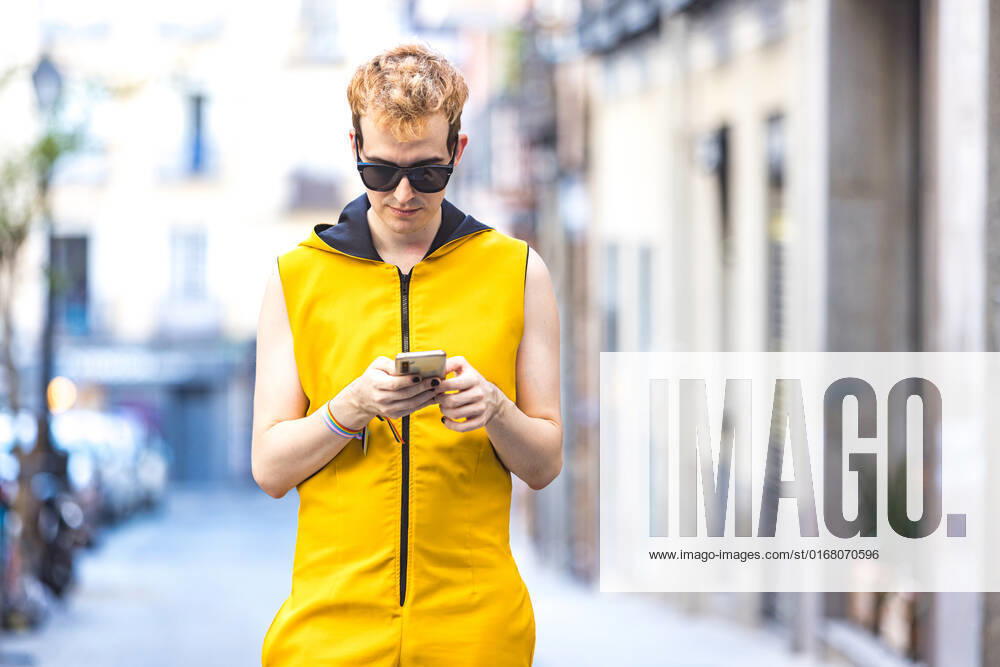 IMAGO Creative, Jahreszeit Sommer, Sommer in der Stadt Cheerful transgender young man in yellow jumpsuit and sunglasses looking at screen while walking and browsing on cellphone on city street DavidBenito_ID643_23435_024