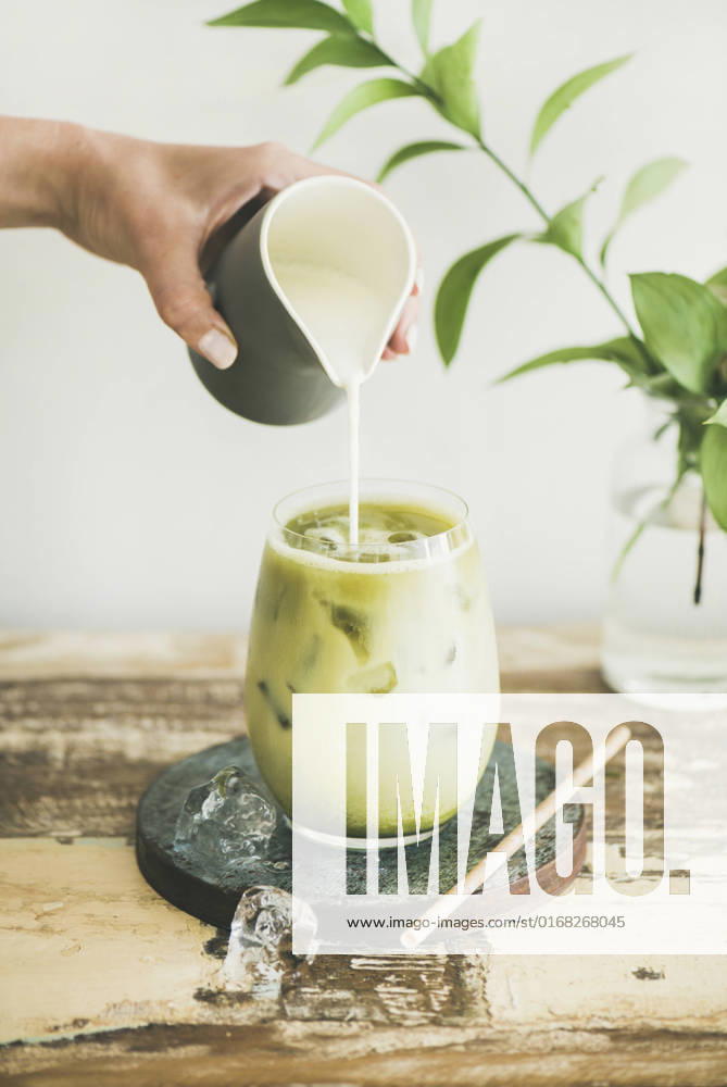 Iced matcha latte drink with milk pouring from pitcher Iced matcha latte drink in glass with coconut milk pouring from pitcher by woman s hand, white wall and plant branches at background. Summer refreshing beverage cold drink