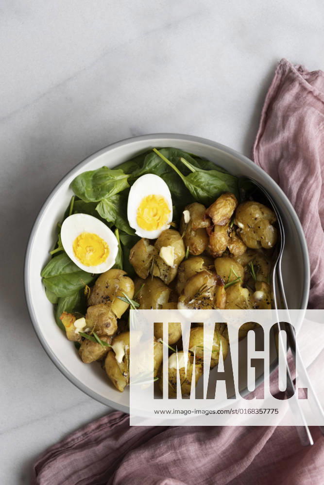 Smashed roasted potatoes in a salad bowl with baby spinach and hard boiled eggs with spoons photographed on a light marble background.