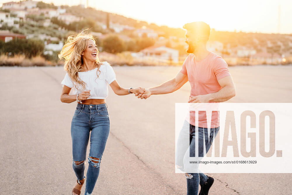 Cheerful man and excited woman looking at each other and laughing while walking dancing holding hands along asphalt road against blurred town on hillside in evening, Model released GustavoPozo_IsaAndKike_97