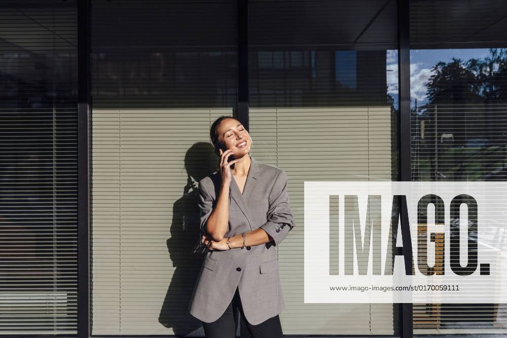 IMAGO Creative: Business, Innovative Technologie-Lösungen Smiling businesswoman with eyes closed talking on mobile phone in front of glass wall model released, Symbolfoto, VPIF07169