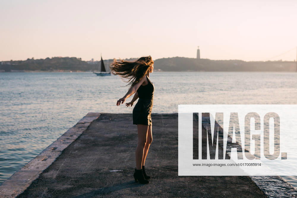 IMAGO Creative, Jahreszeit Sommer, Sommer in der Stadt Attractive charming happy lady in black wear and boots dancing on embankment near water surface with ships in sunny day AlexandraCoelho_PriscillaMarques_12