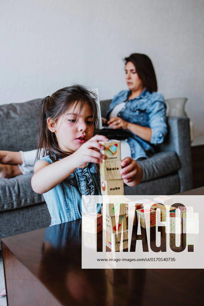 IMAGO Creative: Business - Work-Life Balance Concentrated young Latin American female freelancer sitting on sofa and working remotely on laptop near cute little daughter playing with wooden cube toys at home, Model released, Property released MarcosCastillo_LatinFamily_27