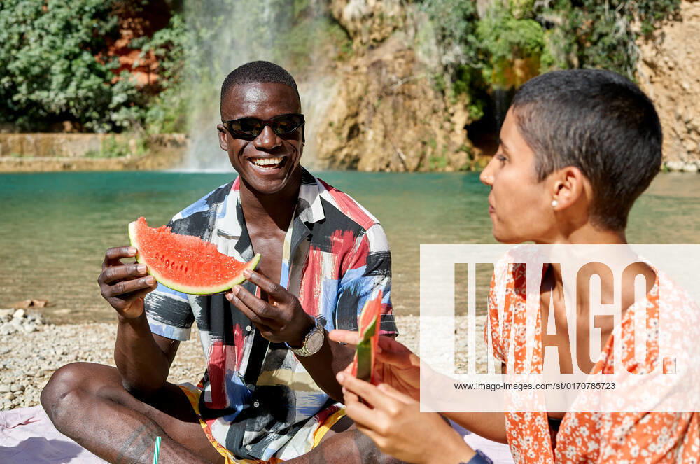 Multiracial cheerful couple sitting together and eating watermelon while having picnic against lake in summer, Model released, Property released AdalbertoRodriguez_ID942_34991_020