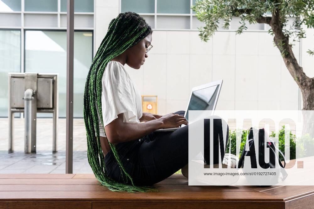 IMAGO Creative: Business - Work-Life Balance Side view of young African American female freelancer with long braided hair and in glasses sitting on wooden bench and browsing laptop in city downtown DavidMunoz_ID994_37337_025