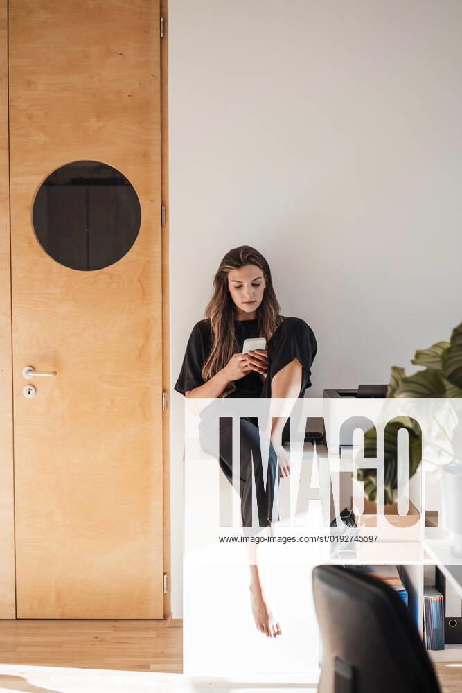 IMAGO Creative: Business, Innovative Technologie-Lösungen Young woman using mobile phone sitting on table at home model released, Symbolfoto property released, JOSEF13433