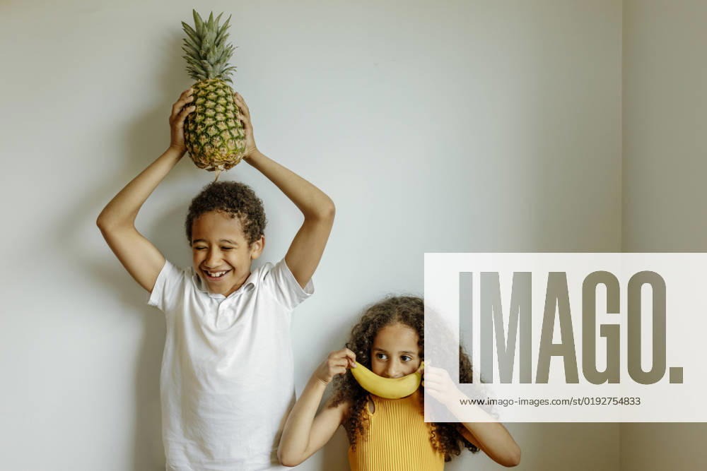 Happy boy holding pineapple above head by sister covering lips with banana in front of wall model released, Symbolfoto property released, VIVF00018