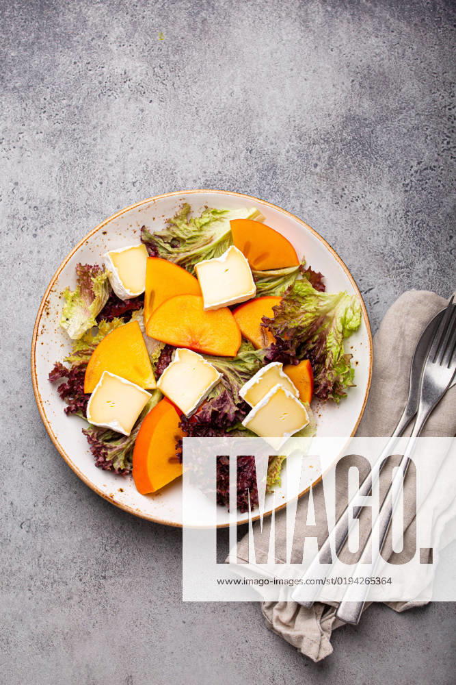 Healthy vegetarian persimmon salad with brie top view Top view flat lay persimmon salad with brie cheese and fresh salad leaves on white plate and stone gray background, seasonal fruit salad as appetizer, vegetarian healthy food