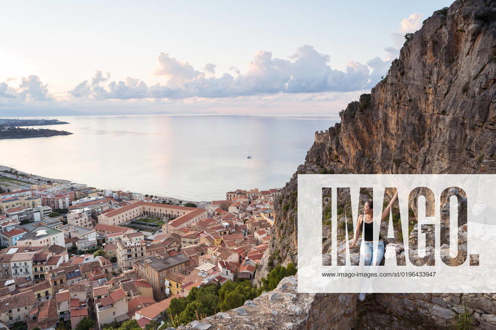 IMAGO Creative, Jahreszeit Sommer, Sommer in der Stadt Woman in casual clothes raising arm and admiring Cefalu City while sitting on old stone wall near mountain slope against sea and cloudy sunset sky in evening in Sicily, Italy JuanAlbertoRuiz_ID10183_308569_012