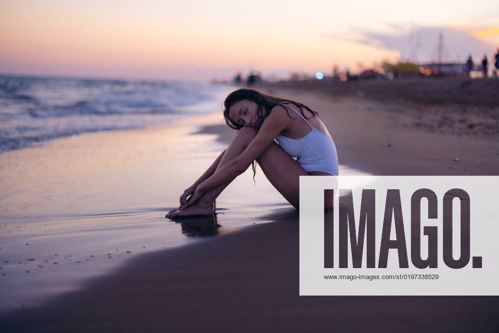 Side view of young woman in white swimwear embracing knees and closing eyes while sitting on beach near sea at sunset, Model released XavierLaiz_ID10743_325282_033