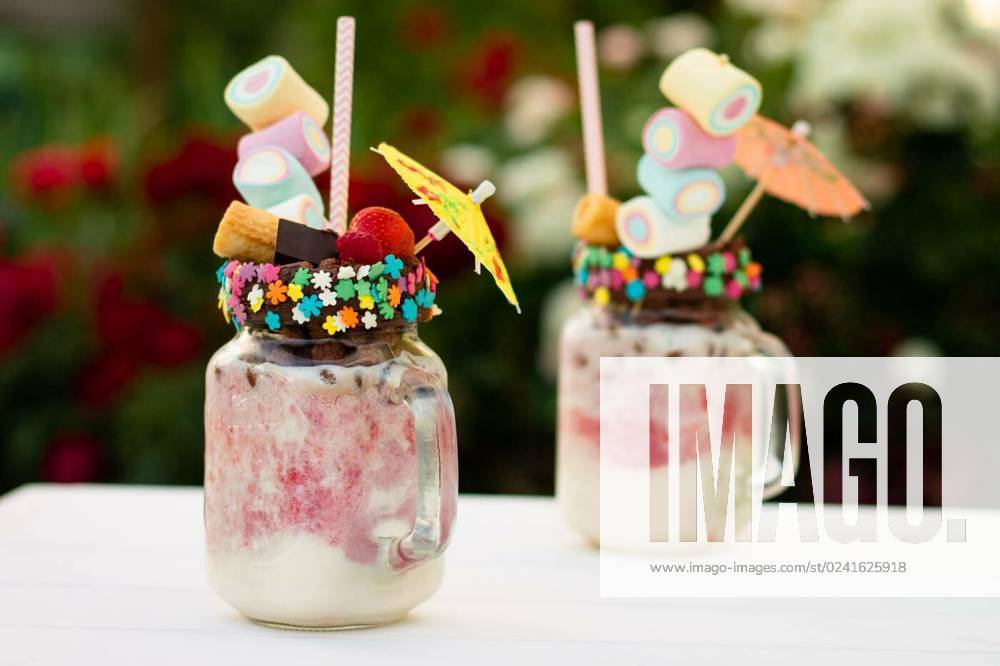 Freakshakes in jars with straw, marshmallow, strawberry, chocolate on a white table on a background of flowers., Freakshakes in jars with cocktail umbrella, straw, marshmallow, strawberry, chocolate on a white table on a background of summer flowers., Freakshakes in jars with cocktail umbrella, straw, marshmallow, strawberry, chocolate on a white table on a background of summer flowers., 26.03.2023,
