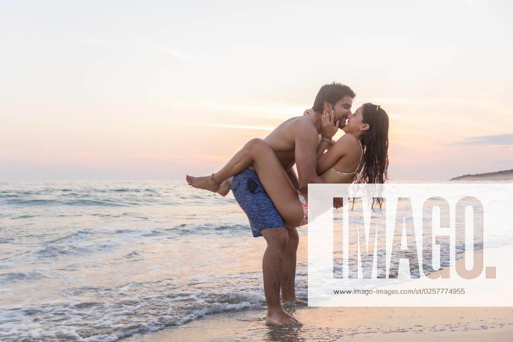 MR_ID13682_13316_EduardoGavira.jpg, MR_ID13682_13317_SamanthaLaguna.jpg Full body of positive young couple in swimwear smiling and embracing while standing on sandy beach near waving sea against sunset sky during summer holidays together in Puerto Escondido, Model released EduardoGavira_ID13682_415222_029