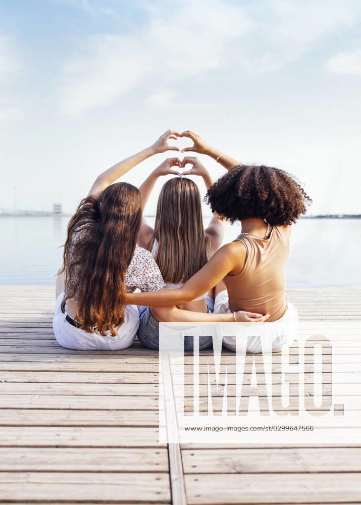 Three female teenagers of different ethnic groups show heart symbols with their hands Three female teenagers of different ethnic groups show heart symbols with their hands. Teen girls in casual clothes sit on wooden bridge near the river or lake and hug. Friendship concept. Copy space ,model released, Symbolfoto